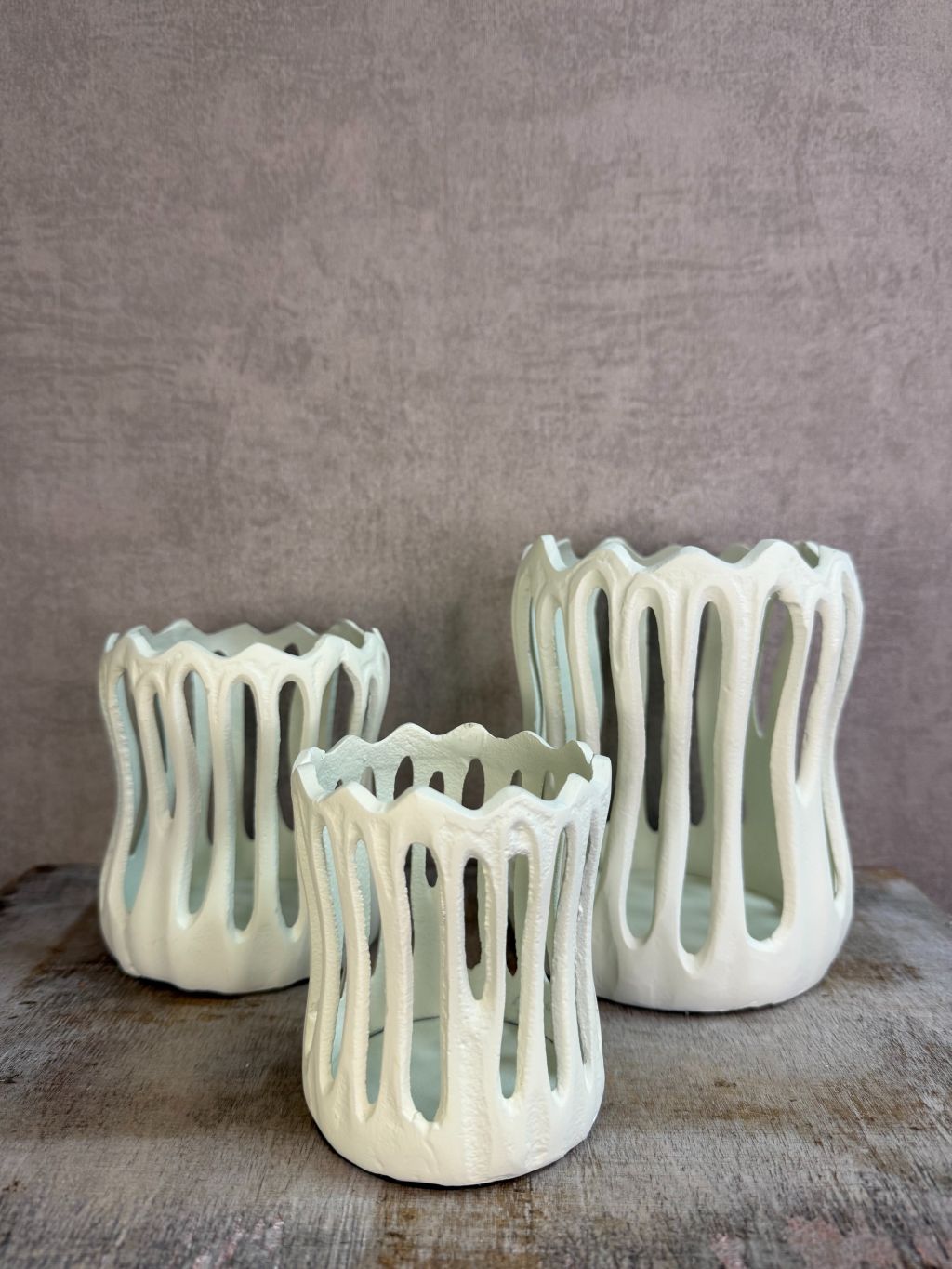 Drie crèmekleurige metalen waxinelichthouders Tivoli in verschillende formaten met open design op een houten ondergrond.