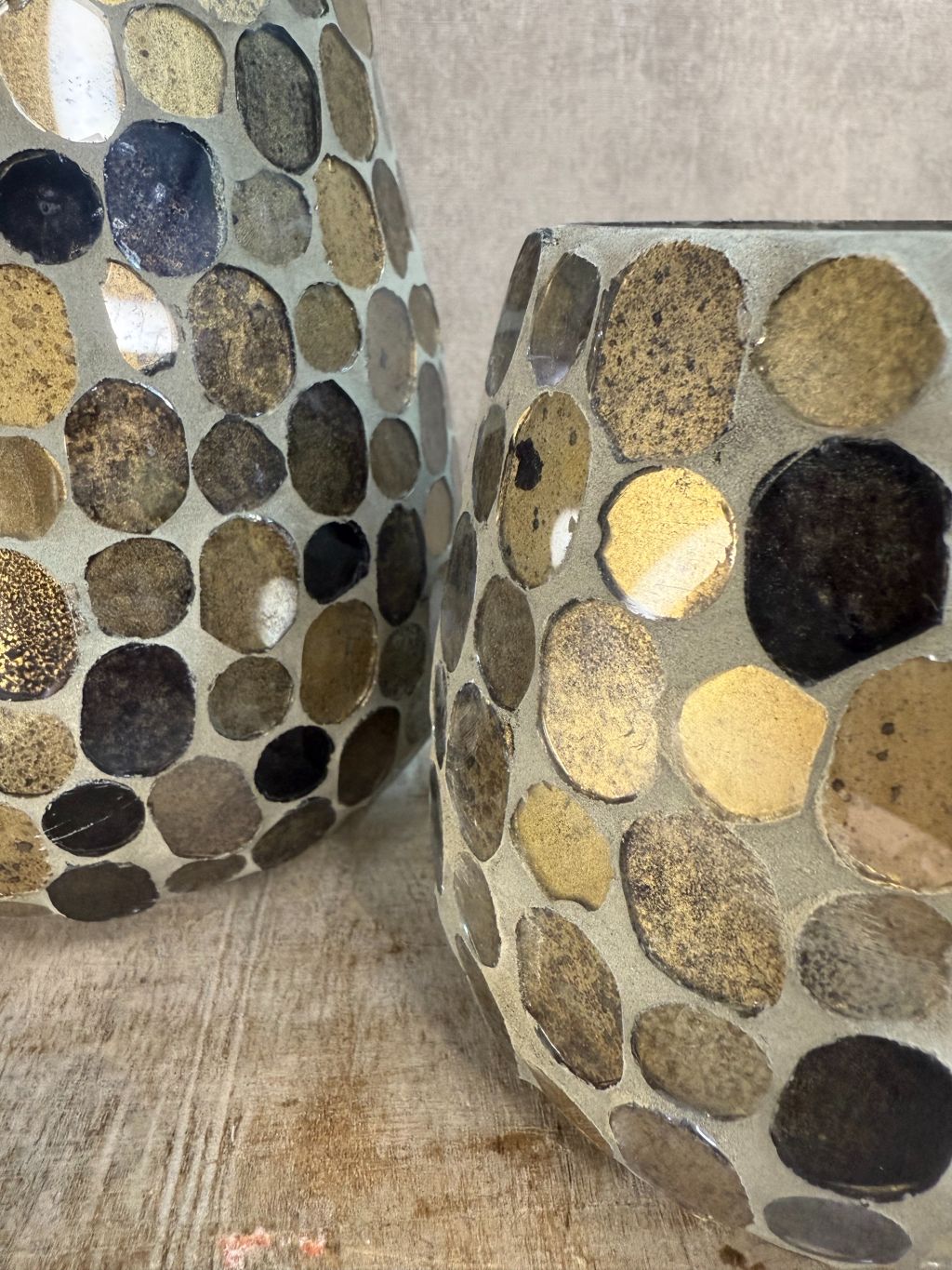 Twee glazen windlichten met mozaïek in aardse tinten van bruin, goud en zwart, staand op een houten ondergrond tegen een grijze muur.