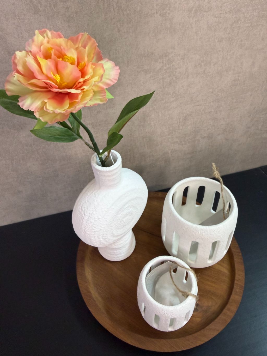 Tafel decoratie set met houten dienblad, witte vaas met bloem en twee witte waxinelichthouders tegen een neutrale achtergrond.